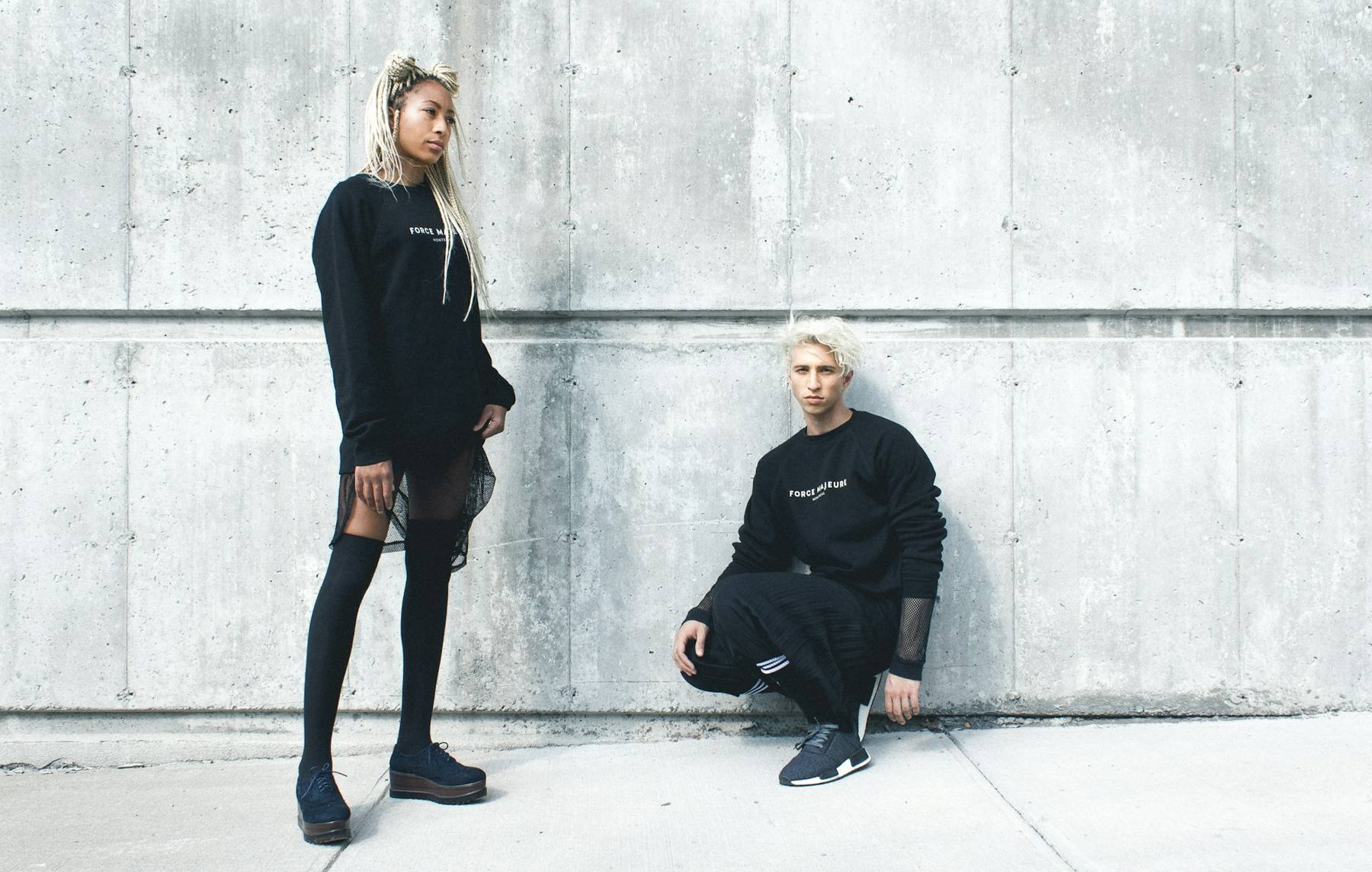 Two people in black streetwear against a concrete wall